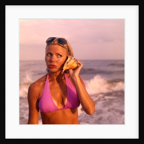 1970s pouting sexy woman in lavender bikini sunglasses near beach surf holding seashell to ear listening to ocean by Anonymous