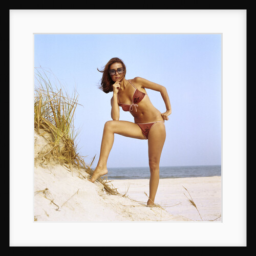 1970s woman wearing two piece string bikini bathing suit sunglasses posing by grassy sand dune at beach by Anonymous
