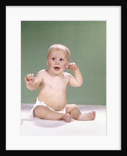 1960s baby wearing diaper sitting on pink blanket mouth open hand touching ear by Anonymous