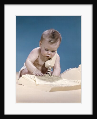 1960s baby reading phone book yellow pages with telephone receiver on shoulder by Anonymous