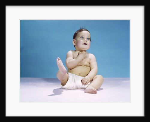 1960s baby wearing diaper sitting looking up and kicking one leg up by Anonymous