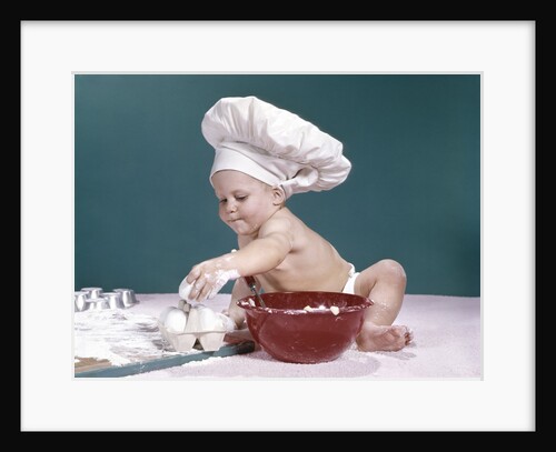 1960s baby wearing chefs hat holding egg surrounded by cooking equipment by Anonymous