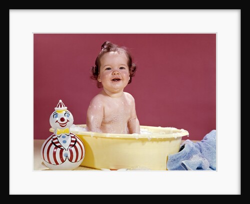1960s smiling brunette baby sitting in yellow plastic bath tub with toy clown looking at camera by Anonymous