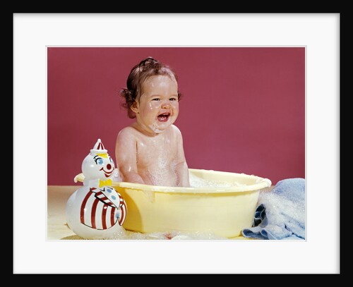 1960s crying brunette baby sitting in yellow plastic bath tub with toy clown by Anonymous