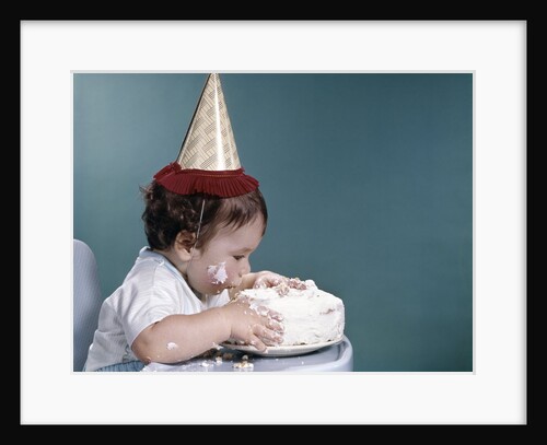 1960s baby in highchair wearing birthday hat eating whole birthday cake by Anonymous