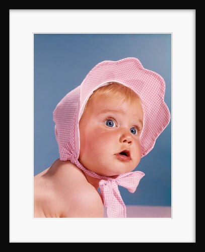 1960s very cute blue eyed baby wearing pink white checked bonnet looking up by Anonymous