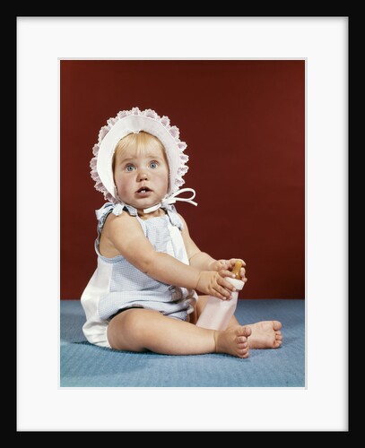 1960s baby girl ruffled bonnet holding bottle funny facial expression looking at camera by Anonymous
