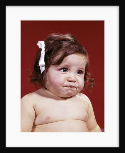 1960s portrait of chubby unhappy baby girl with white ribbon bow in messy hair by Anonymous