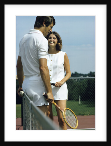 1970s smiling couple standing by net on tennis court holding wood rackets by Anonymous