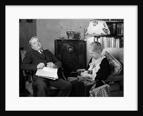 1930s elderly couple sitting in living room listening to radio woman knitting man with newspaper in lap by Anonymous