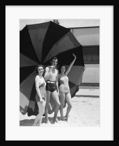 1930s two women one man smiling wearing bathing suits standing under extra large striped beach umbrella by Anonymous