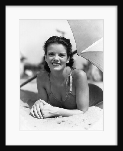 1930s teen girl lying on beach under umbrella wearing bathing suit smiling looking at camera by Anonymous