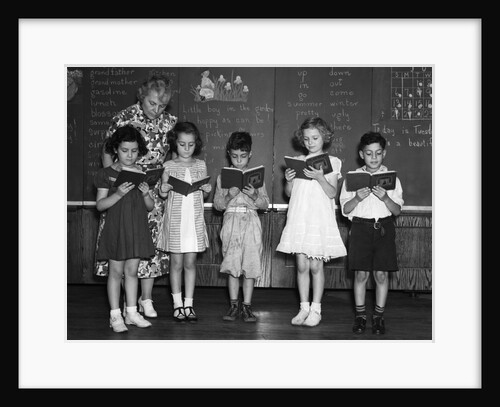 1930s line-up of 5 elementary school students in front of blackboard reading books with teacher looking on by Anonymous