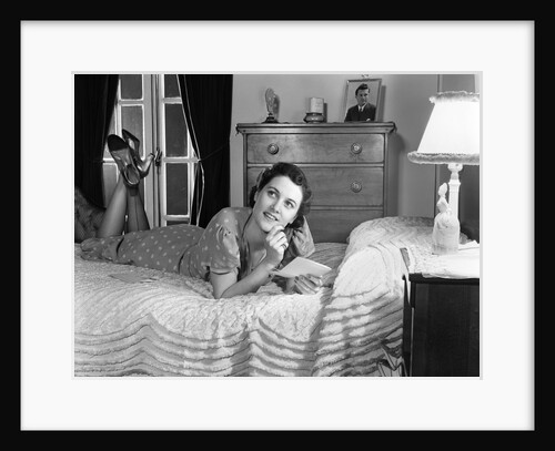 1940s fully dressed smiling woman laying on her bed with her legs up reading a letter in her bedroom by Anonymous