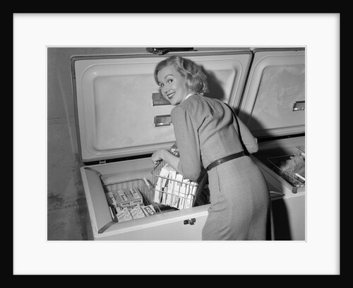 1950s blond woman lifting wire basket food items from a deep freezer looking at camera over her shoulder by Anonymous