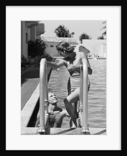 1930s woman wearing one piece bathing suit climbing out of swimming pool looking down at man smiling by Anonymous