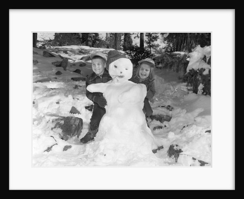 1950s 1960s smiling boy and girl building a snowman together in snowy woods by Anonymous