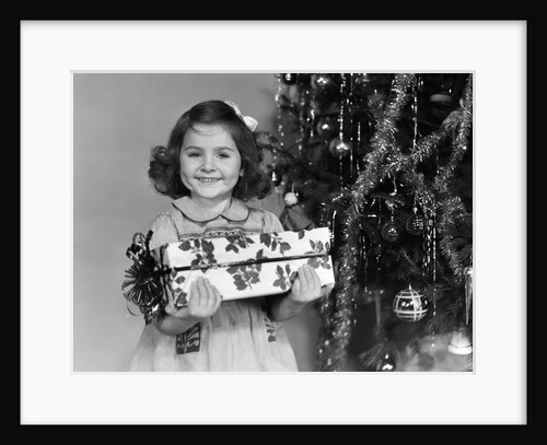 1940s little girl standing near christmas tree holding a wrapped present smiling looking at camera by Anonymous