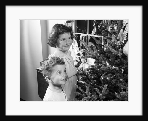 1950s boy girl wearing pajamas smiling up at christmas tree decorations by window by Anonymous