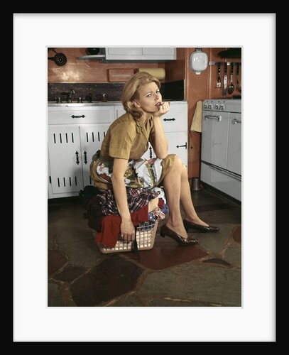 1960s Tired Housewife Sitting On Top Of Laundry Basket In Kitchen by Anonymous