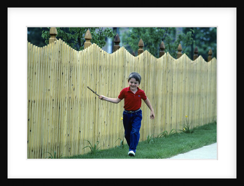 1980s Smiling Boy Running Along Sidewalk Rattling Stick On Tall Picket Fence Making Noise by Anonymous