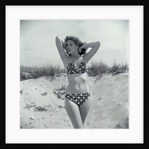 1950s Brunette Bathing Beauty In Polka Dot Bikini Standing In Sand With Hands Behind Head Looking At Camera by Anonymous