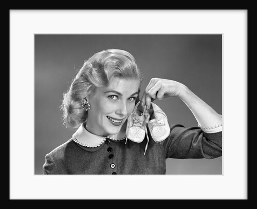 1950s 1960s Smiling Blond Woman Communicating That She Is Mother To Be By Holding Up A Pair Of Baby Shoes Looking At Camera by Anonymous