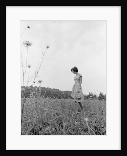 1970s Woman Wearing Dress Carrying Large Straw Hat Standing In Middle Of Field Looking Down by Anonymous