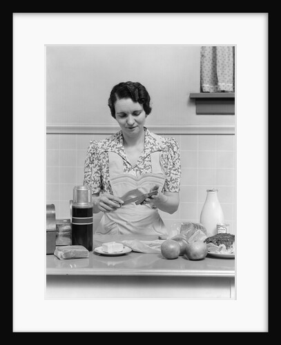1930s Woman Housewife In Kitchen Wearing Apron Making Sandwich Packing Metal Lunch Box Thermos Apples Bread Milk On Counter by Anonymous