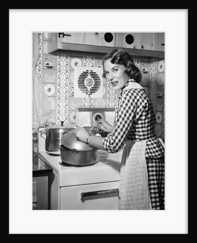 1950s Housewife Wearing Checkered Dress Standing In Kitchen Stirring Pot On Stove Looking Over Shoulder by Anonymous