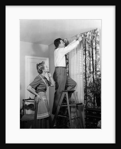 1950s Skeptical Wife In Apron Fingers Pressing Chin Watching Husband On Ladder Hanging New Jungle Motif Drapes In Living Room by Anonymous