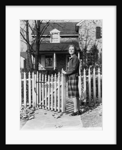 1940s Smiling Pretty Young Teenage Girl Standing By White Picket Fence In Front Of Stone House In Autumn Looking At Camera by Anonymous