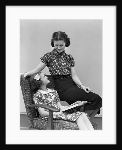 1930s Smiling Little Girl Sitting In Chair Reading Looking Up At Woman Older Sister Sitting On Arm Of The Chair by Anonymous