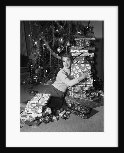 1950s portrait of young woman holding stack of presents next to a Christmas tree by Anonymous