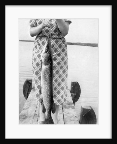 Woman holds on large Northern Pike on a lake pier, ca. 1950. by Anonymous