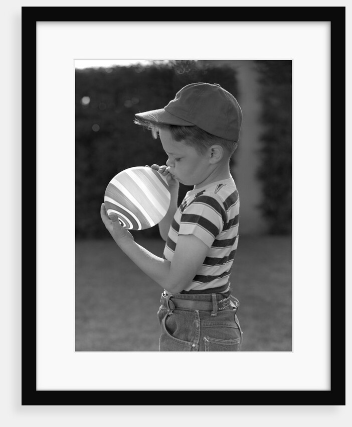 1950s Side View Of Boy In Baseball Cap and Striped T-Shirt Blowing Up Swirled Balloon by Anonymous