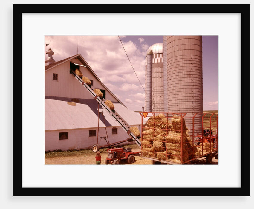 1970s Girl Watching Hay Bales On Conveyor Belt Loading Into Barn By Farm Grain Silos by Anonymous