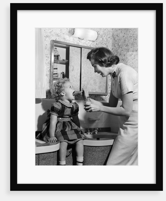1950s Mother Feeding Medicine To Young Daughter With Mouth Wide Open Sitting On Bathroom Sink by Anonymous