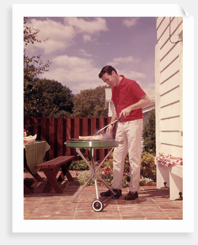 1960s Man Wearing Red Shirt Cooking Steak Outdoor On Backyard Grill by Anonymous