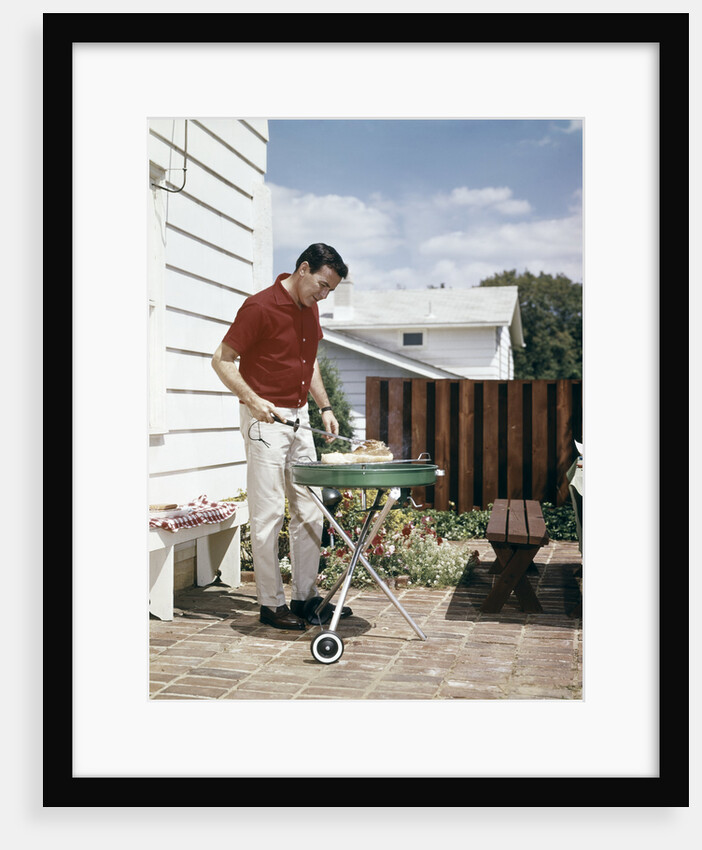 1960s Man Wearing Red Shirt Grilling Steak On Backyard Brick Patio by Anonymous