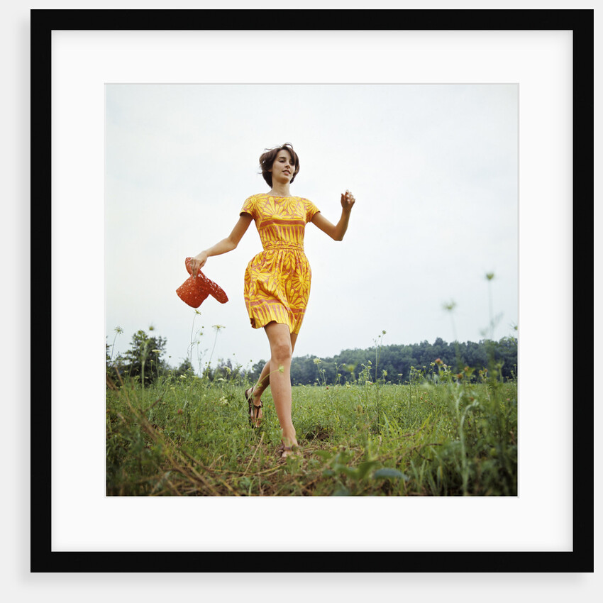 1970s Young Woman Wearing Colorful Print Dress Running In Field Carrying Red Straw Hat by Anonymous