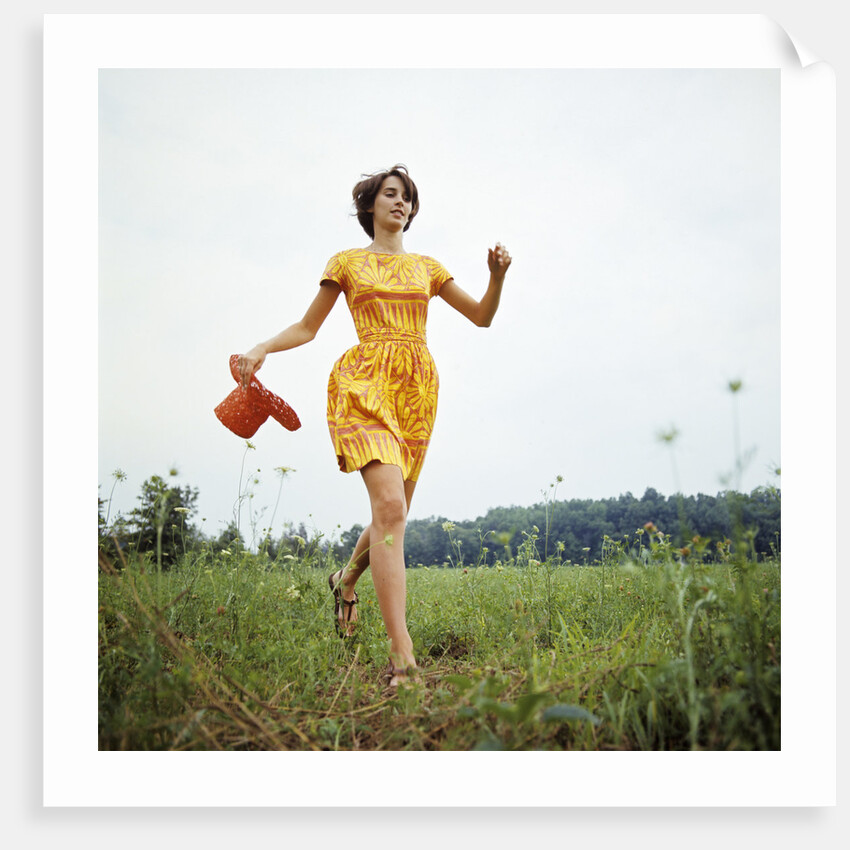 1970s Young Woman Wearing Colorful Print Dress Running In Field Carrying Red Straw Hat by Anonymous