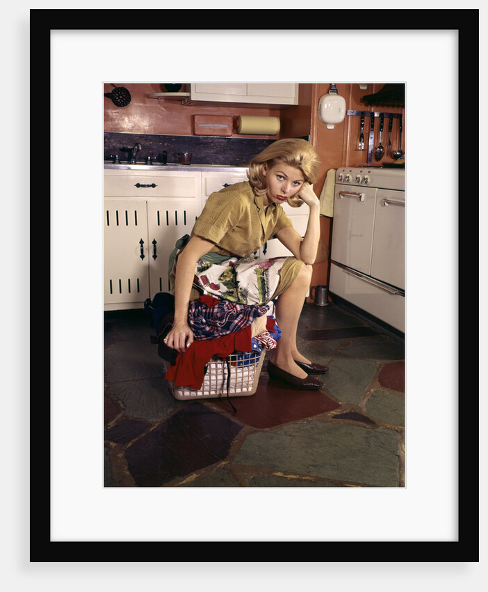 1960s Weary Dejected Woman Housewife Homemaker Sitting On Full Laundry Basket In Kitchen by Anonymous