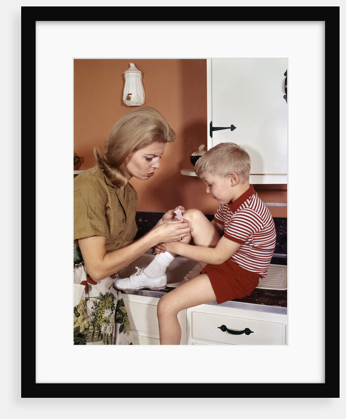 1970s Mother Giving First-Aid Putting Band-Aid On Knee Of Son In Kitchen by Anonymous