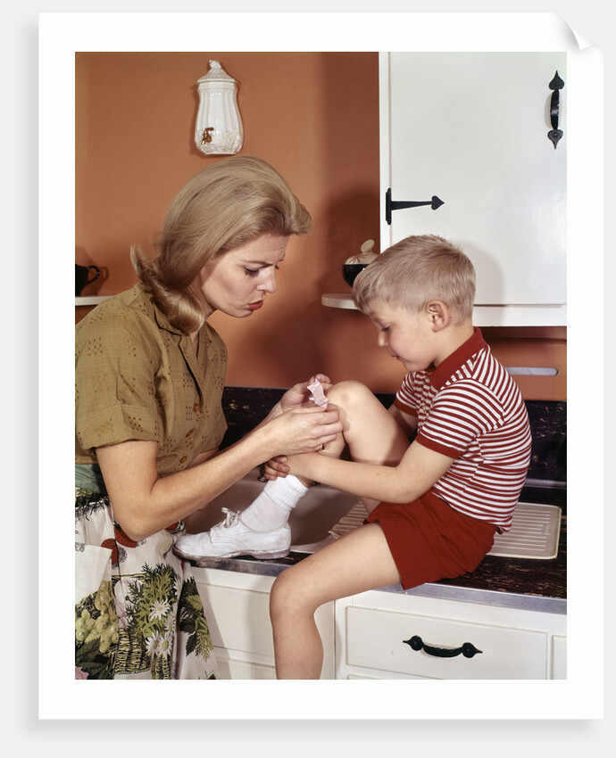 1970s Mother Giving First-Aid Putting Band-Aid On Knee Of Son In Kitchen by Anonymous