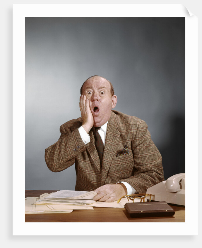 1960s Shocked Balding Executive With Hand To Cheek Sitting At Desk by Anonymous