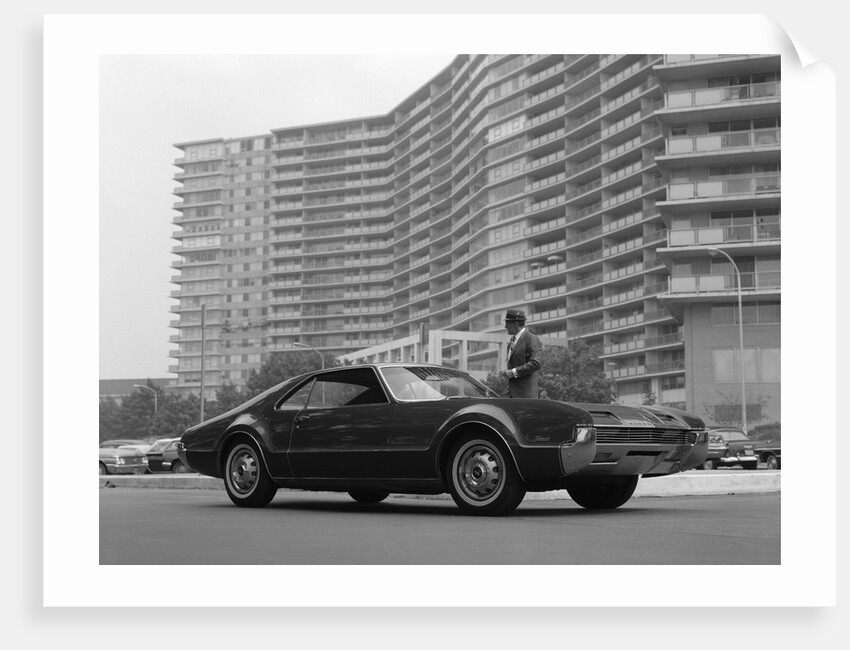 1960s Man In Suit and Hat Standing Beside Oldsmobile Toranado In Front Of Large Apartment Building by Anonymous
