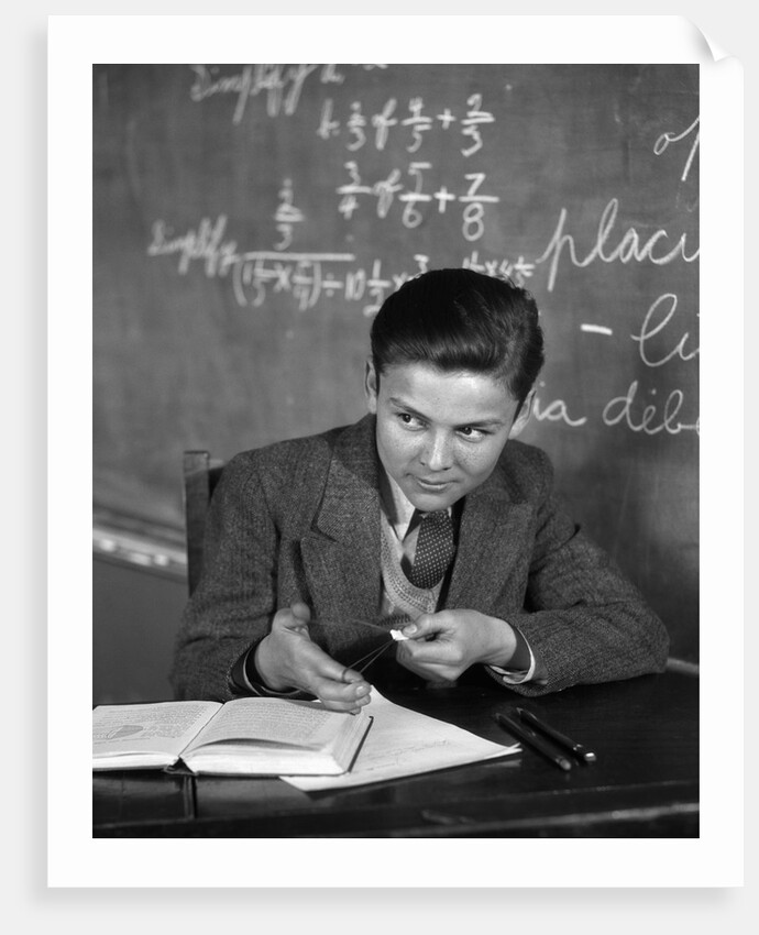 1920s 1930s Boy At Desk In Classroom In Front Of Blackboard Shooting Paper Wad With Rubber Band by Anonymous
