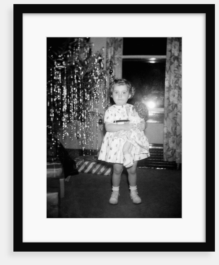 A little girl stands by the Christmas tree clutching her doll, ca. 1955 by Anonymous