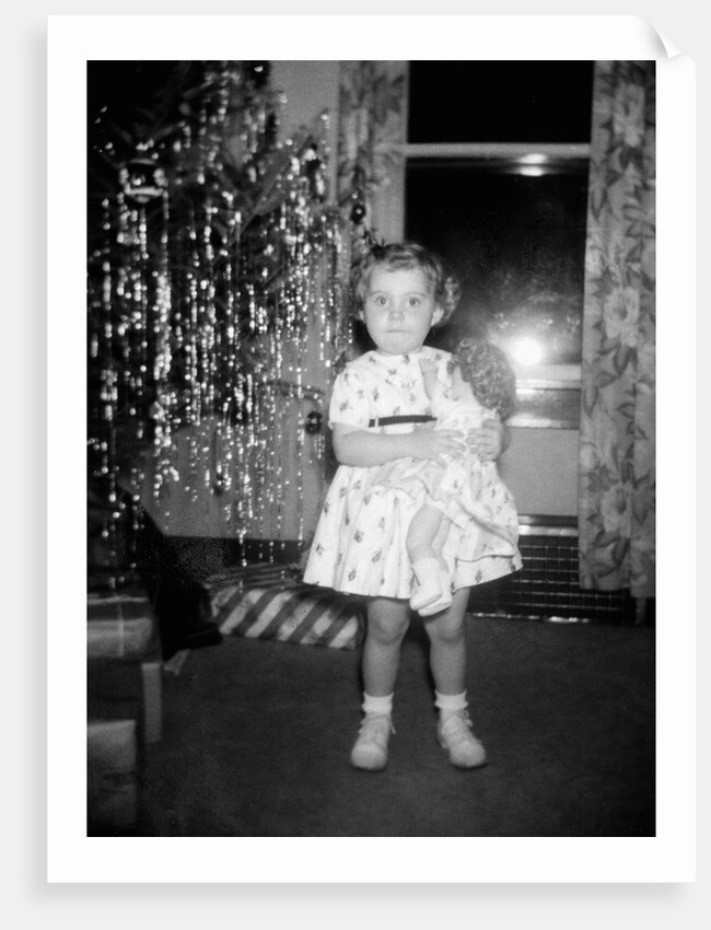 A little girl stands by the Christmas tree clutching her doll, ca. 1955 by Anonymous
